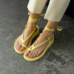 May include: Yellow sandals with a toe loop and ankle straps. The sandals have a flat sole and are made of a soft material. The ankle straps are tied in a bow. The sandals are shown on a gray surface.