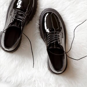 May include: A pair of black patent leather oxford shoes with a high-gloss finish, rounded toes, and thick, lugged rubber soles. The shoes feature matching black laces that are tied, with a low-cut silhouette and a structured, polished appearance.