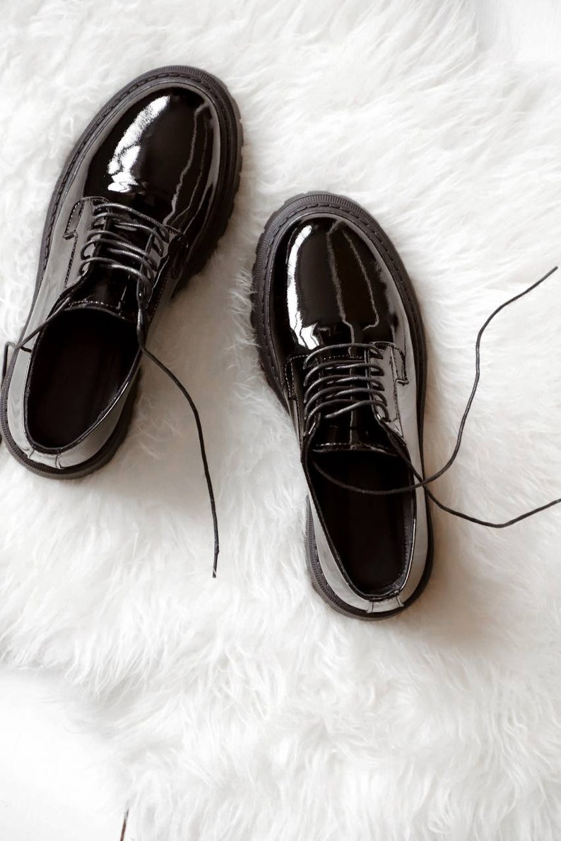May include: A pair of black patent leather oxford shoes with a high-gloss finish, rounded toes, and thick, lugged rubber soles. The shoes feature matching black laces that are tied, with a low-cut silhouette and a structured, polished appearance.