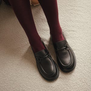 May include: A pair of black leather loafers with a decorative metal buckle. The shoes are on a light gray carpet, and are paired with burgundy tights. The loafers have a rounded toe and a low heel.