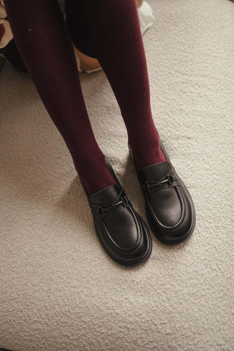 May include: A pair of black leather loafers with a decorative metal buckle. The shoes are on a light gray carpet, and are paired with burgundy tights. The loafers have a rounded toe and a low heel.