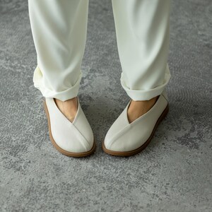 May include: A pair of white, perforated slip-on shoes with a V-shaped vamp and a light brown sole. The shoes are paired with cream-coloured trousers with the cuffs rolled up. The shoes appear to be made of a breathable material.