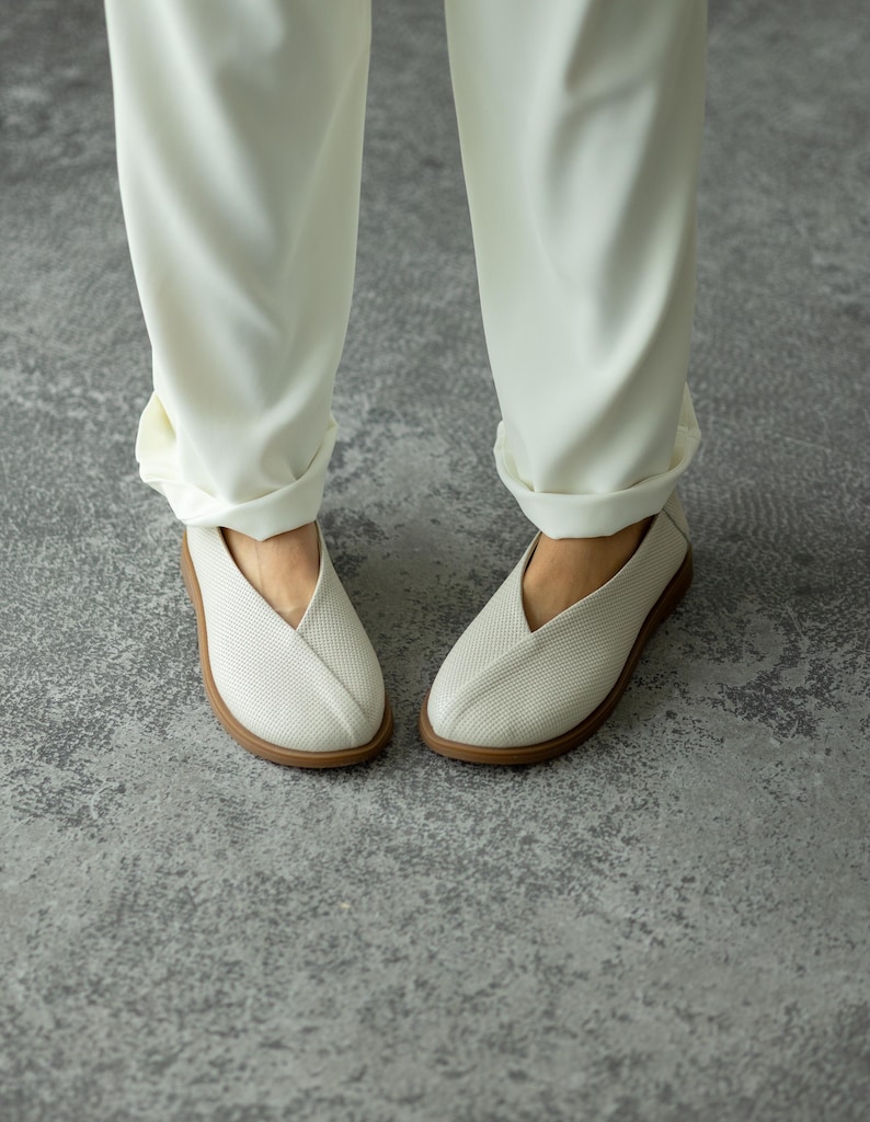May include: A pair of white, perforated slip-on shoes with a V-shaped vamp and a light brown sole. The shoes are paired with cream-coloured trousers with the cuffs rolled up. The shoes appear to be made of a breathable material.