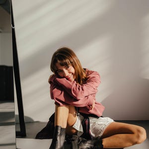 May include: A woman wearing a pink sweatshirt and white shorts sits on a white floor with her legs crossed. She is wearing black ankle boots and smiling at the camera.
