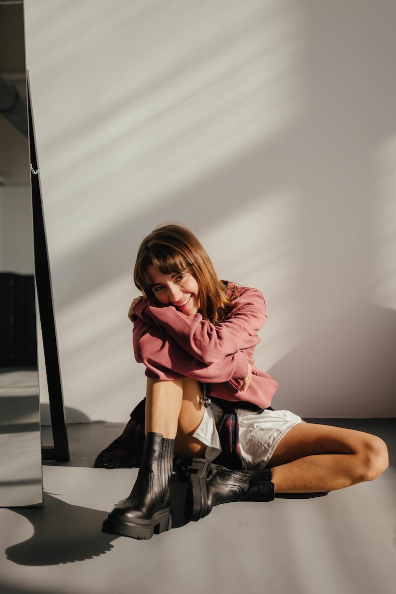 May include: A woman wearing a pink sweatshirt and white shorts sits on a white floor with her legs crossed. She is wearing black ankle boots and smiling at the camera.