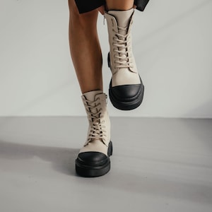 May include: A pair of beige leather lace-up boots with black rubber soles. The boots have a chunky platform heel and are worn by a person standing on a light gray surface.