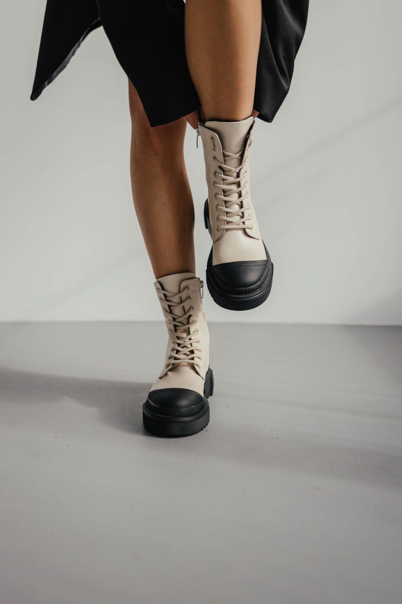 May include: A pair of beige leather lace-up boots with black rubber soles. The boots have a chunky platform heel and are worn by a person standing on a light gray surface.