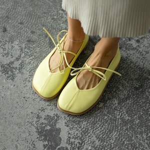 Puede incluir: Bailarinas amarillo pálido con punta cuadrada y cierre de lazo. Los zapatos son de material liso y tienen suela marrón claro. Están sobre una superficie de hormigón gris.