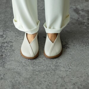 May include: A pair of white, textured slip-on shoes with a distinctive V-shaped design. The shoes have a brown sole and are worn with white, cuffed trousers. The image is taken from a low angle, showcasing the footwear and the lower portion of the trousers.