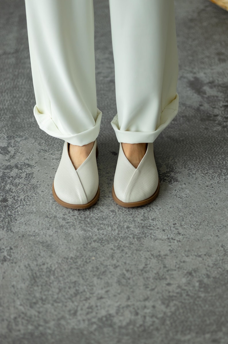 May include: A pair of white, textured slip-on shoes with a distinctive V-shaped design. The shoes have a brown sole and are worn with white, cuffed trousers. The image is taken from a low angle, showcasing the footwear and the lower portion of the trousers.