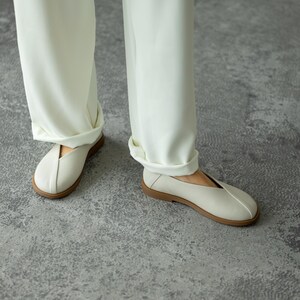 May include: Cream-coloured slip-on shoes with a textured upper and a brown sole. The shoes are paired with white trousers. The shoes appear to be made of a breathable material, suitable for casual wear.