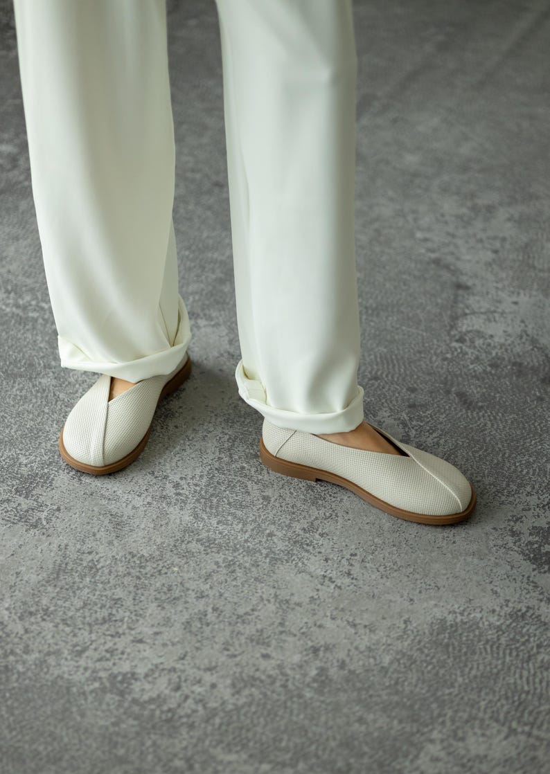 May include: Cream-coloured slip-on shoes with a textured upper and a brown sole. The shoes are paired with white trousers. The shoes appear to be made of a breathable material, suitable for casual wear.