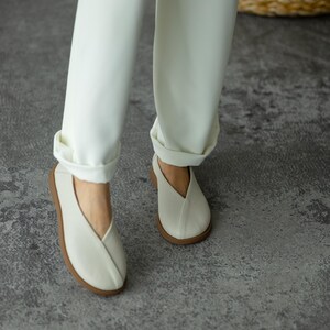 May include: Off-white slip-on shoes with a V-shaped vamp and a brown sole. The shoes are worn with white trousers with turned-up hems. The shoes appear to be made of a textured material.