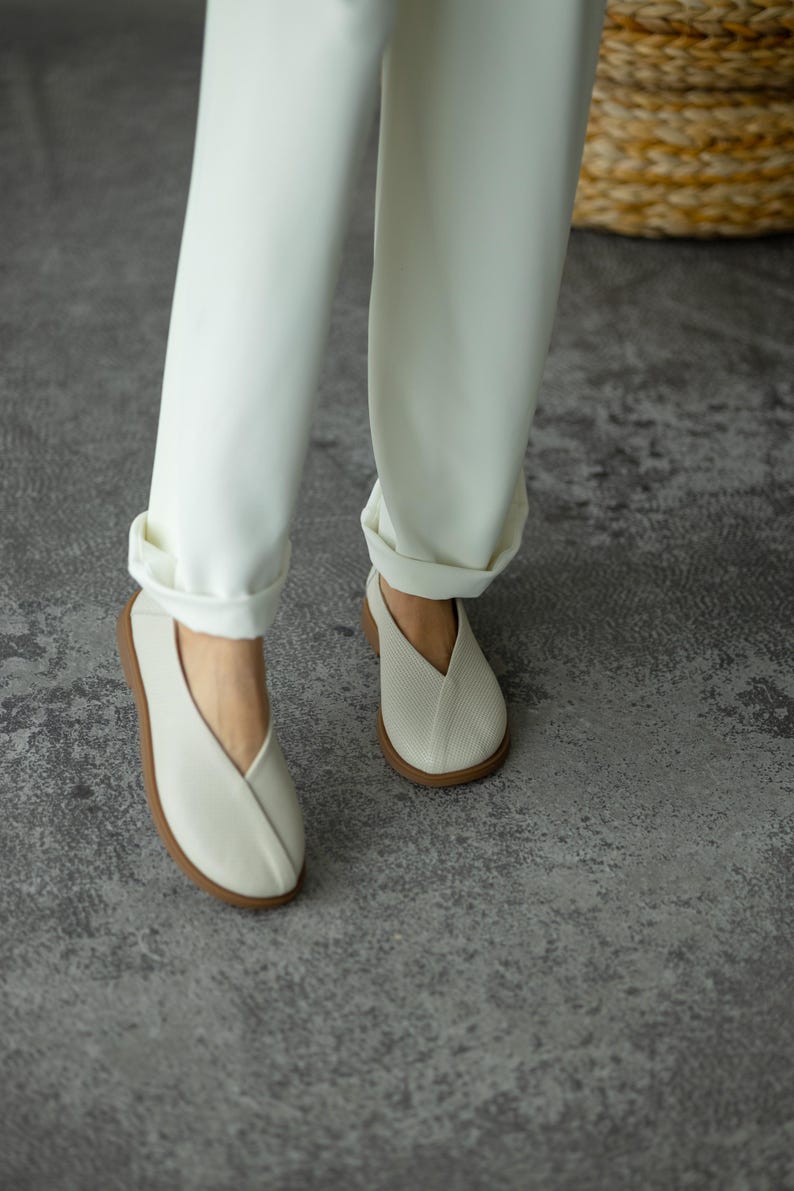 May include: Off-white slip-on shoes with a V-shaped vamp and a brown sole. The shoes are worn with white trousers with turned-up hems. The shoes appear to be made of a textured material.