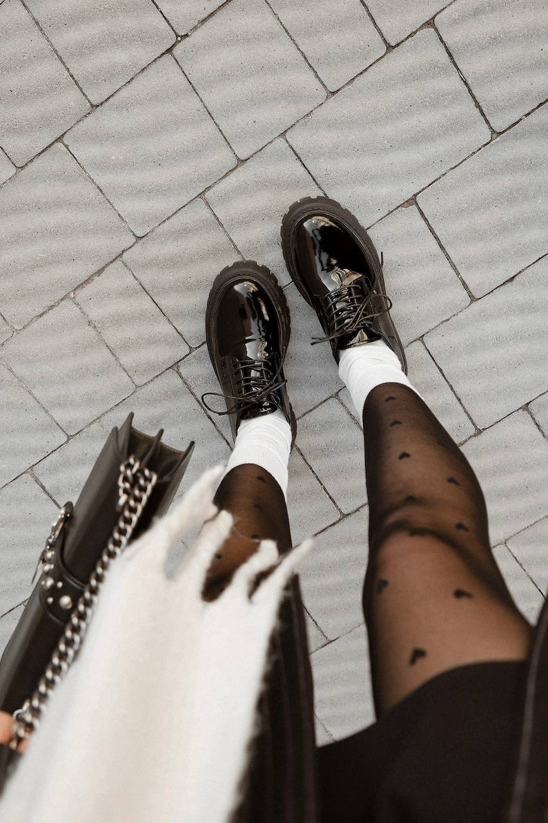 May include: A pair of black patent leather lace-up shoes with thick lug soles, worn with white crew socks and sheer black tights featuring a small heart pattern. A black handbag with a silver chain strap is visible in the lower left corner.