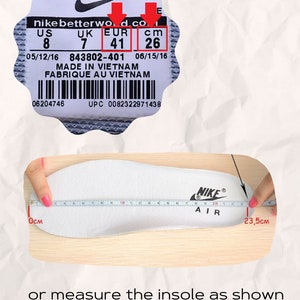 May include: A white Nike Air shoe insole with a black Nike logo and the text "Air" printed on it. The insole is being measured with a tape measure, showing a measurement of 23.5 cm. The image also shows a label with the text "nike betterworld.co.e" and size information: US 8, UK 7, EUR 41, cm 26. The label also includes the text "Made in Vietnam, Fabrique au Vietnam", a date code, and a UPC code.