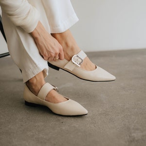 Puede incluir: Un par de zapatos de cuero de color blanco roto con punta puntiaguda y una correa con hebilla. Los zapatos tienen un tacón bajo y los lleva una persona con pantalones blancos.