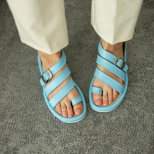 May include: Light blue sandals with multiple straps and silver buckles. The sandals have a toe loop and are worn with cream-colored pants. The footwear is on a gray surface.