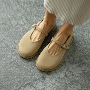 Puede incluir: Zapatos Mary Jane de ante color beige con punta redondeada y correa con hebilla lateral. Los zapatos tienen suela marrón claro y se muestran sobre una superficie gris.