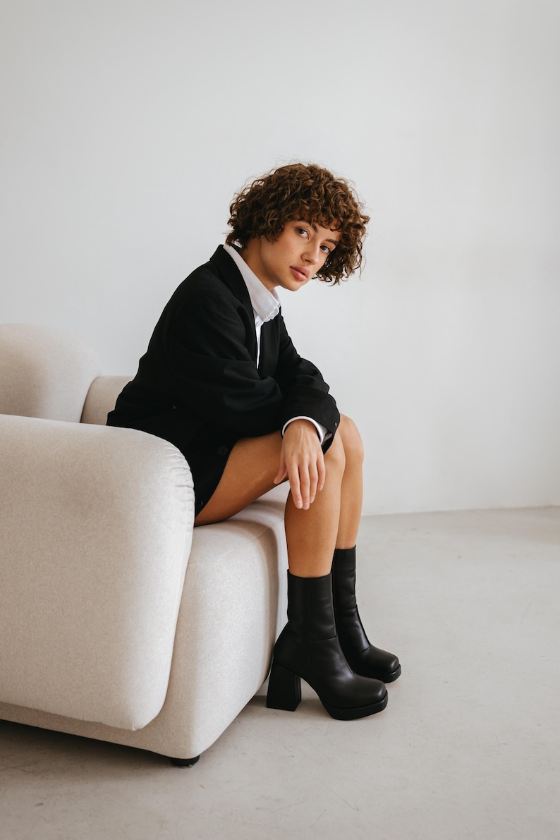 May include: A person wearing a black blazer over a white collared shirt, paired with black platform boots. The person is seated on a beige, modern-style sofa. The background is a plain white wall.