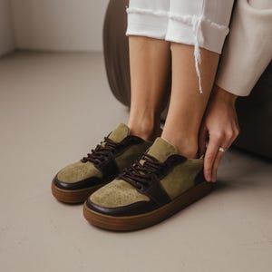 Person wearing khaki leather and olive suede wide toe sneakers, sitting on a chair with white pants, adjusting the shoe, minimalist background