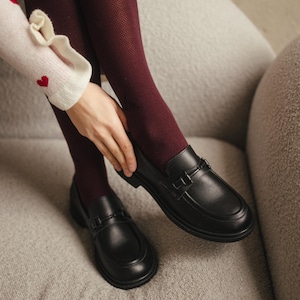 May include: Black leather loafers with a decorative strap buckle. The shoes are on a gray surface, paired with burgundy tights. The image shows a close-up of the shoes and lower legs.