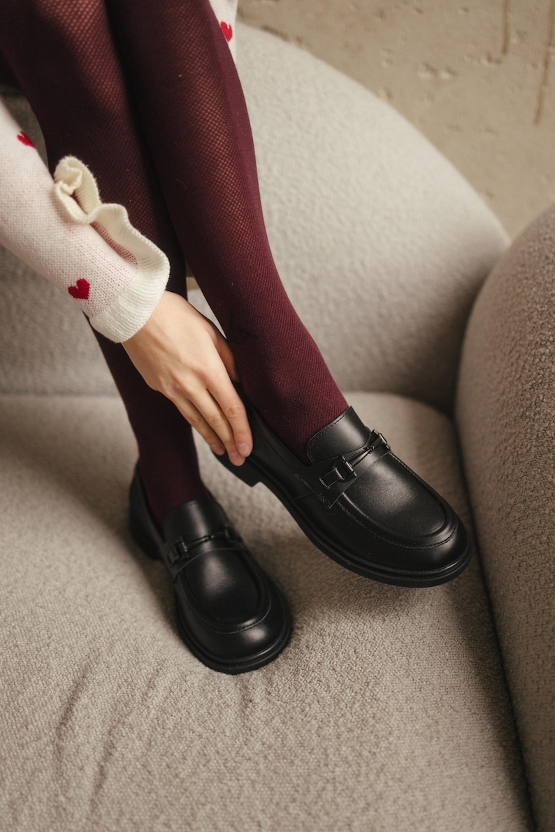 May include: Black leather loafers with a decorative strap buckle. The shoes are on a gray surface, paired with burgundy tights. The image shows a close-up of the shoes and lower legs.