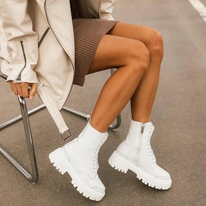 Puede incluir: Botas blancas de cuero con cordones y suela de plataforma gruesa. Las botas tienen una cremallera lateral y las lleva puestas una persona sentada en una silla.