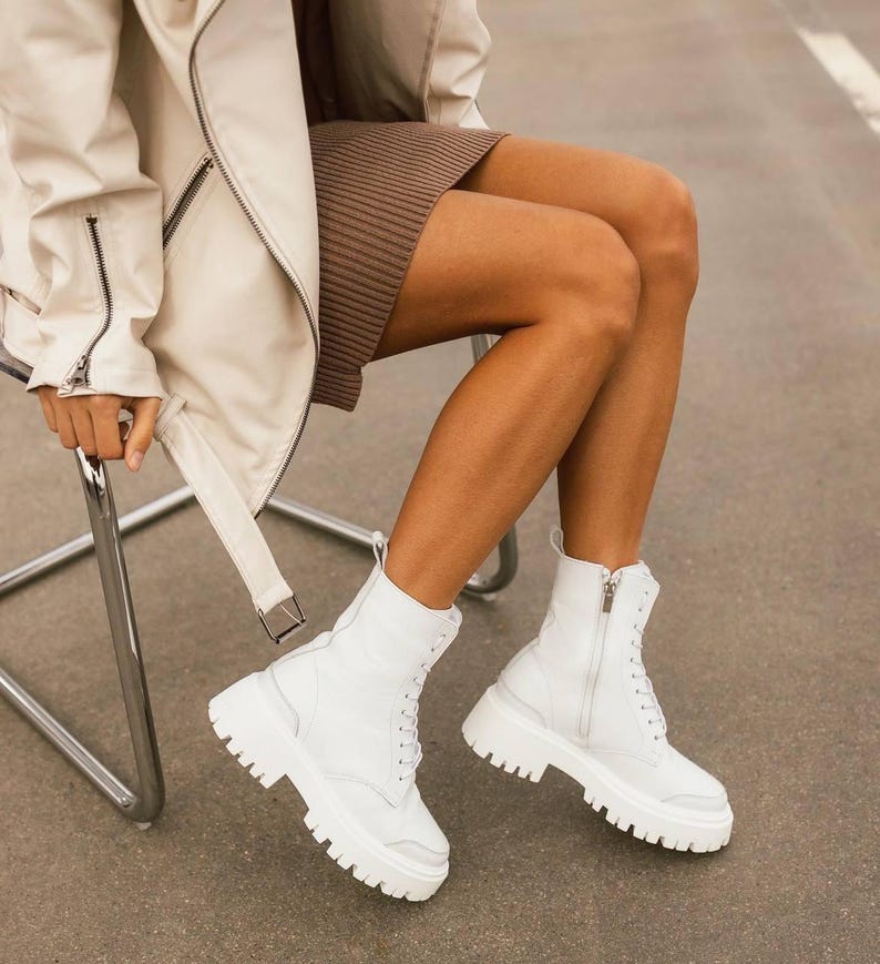 Puede incluir: Botas blancas de cuero con cordones y suela de plataforma gruesa. Las botas tienen una cremallera lateral y las lleva puestas una persona sentada en una silla.