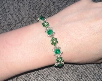 May include: A beaded bracelet featuring a series of green flower-shaped beads interspersed with silver-colored beads. The bracelet is worn on a wrist, showcasing the jewelry's design and color contrast. The bracelet is a piece of handmade jewelry.