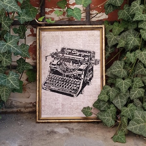May include: A framed print featuring a black and white illustration of an Olympia typewriter. The print is set against a background of dictionary pages and framed in a gold-colored frame. The image is surrounded by green ivy.