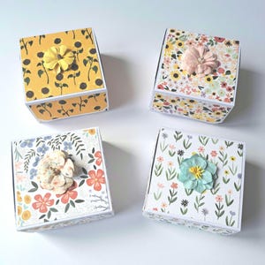 May include: Four small square boxes with floral patterns and a single flower embellishment on each lid. The boxes are yellow, pink, white, and blue.