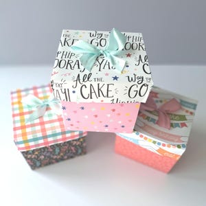 May include: Three small gift boxes with colourful patterned paper. The top box is white with black text that says "Way to go" and "All the cake". The middle box is pink with white stars and the bottom box is red with white text that says "Happy birthday" and "Eat cake".