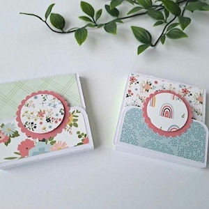 May include: Two decorative paper card holders with floral and rainbow designs. Each holder features a scalloped edge circle with a floral or rainbow pattern. The holders have a white base and are decorated with pastel colours.