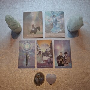 May include: A collection of tarot cards, crystals, and stones arranged on a light-colored surface. The cards feature illustrations and text, including "Believing is Seeing" and "The Lovers." Two large, light-colored stones flank the cards.
