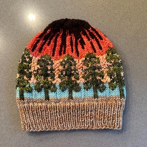 May include: A knitted hat with a colorful design featuring a sunset over a forest. The hat is brown, orange, green, blue, and black.