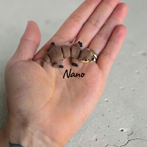 May include: A small, articulated brown otter figurine with black paws and a cream-colored face, featuring the word Nano printed in black script text below the figure. The otter has a segmented body design and dark eyes.