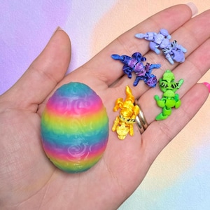 May include: A rainbow-colored egg with a textured swirl pattern alongside four small, articulated dragon figurines in light blue, dark blue, yellow, and green. Each dragon features black painted eyes and segmented bodies for movement.