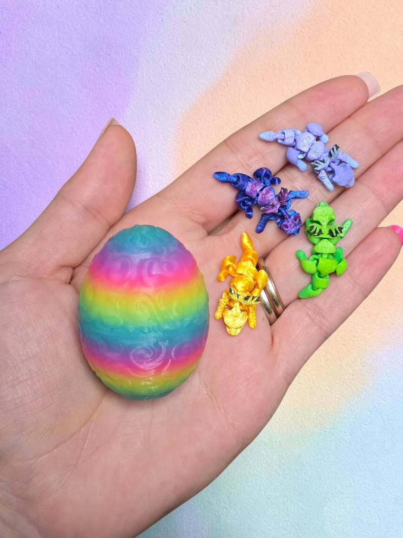 May include: A rainbow-colored egg with a textured swirl pattern alongside four small, articulated dragon figurines in light blue, dark blue, yellow, and green. Each dragon features black painted eyes and segmented bodies for movement.