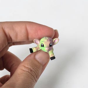 May include: A miniature lamb figurine in shades of green, pink, and yellow. The small lamb has black hooves and large, dark eyes. The figurine is held between fingers against a white background.