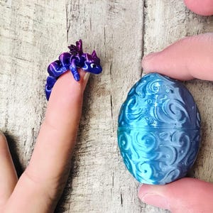 May include: A small blue and purple dragon ring is on a finger, beside a blue egg-shaped container with a swirling design. The dragon has detailed wings and a curled body. The egg is held in a hand.