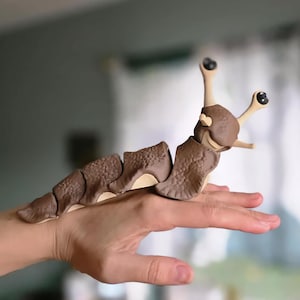 Könnte beinhalten: Ein braunes und beiges, bewegliches Spielzeug-Tausendfüßler mit großen, schwarzspitzigen Antennen. Der segmentierte Körper wird in einer Hand gehalten, was das Design und die Farbgebung des Spielzeugs zeigt. Das Spielzeug besteht aus einem harten Material.