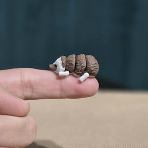 Könnte beinhalten: Eine Miniatur-Igel-Figur mit braunem und weißem Körper, die auf einer Fingerspitze ruht. Die kleine Skulptur hat eine detaillierte Textur und liegt. Die Figur ist ein bezaubernder Dekorationsartikel.