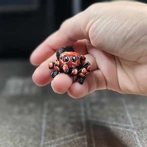 以下が含まれることがあります： 小さなクモのフィギュア。赤い体に黒い模様、8本の黒い脚、オレンジ色の目があります。手で持っています。詳細なモデルです。