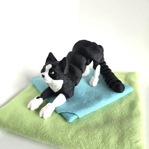 May include: A black and white cat figurine in a stretching pose, with white paws and a white face with green eyes. The cat is lying on a blue and green fabric surface.