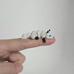 May include: A small, white and black fox figurine. The fox is lying down, with its body made of connected white pieces. The fox has black ears and feet. The figurine is resting on a finger.