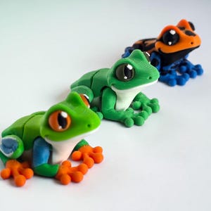 May include: Three colorful frog figurines. The frogs are green, orange, blue, and black. The frogs have large eyes and are posed as if they are sitting. The figurines are on a white surface.