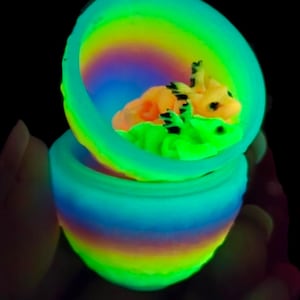 May include: A glowing, multi-colored, rainbow-striped egg-shaped container that is open at the top. Inside the container, two small, detailed, light-colored creatures with dark markings are nestled together, illuminated by a vibrant neon light.