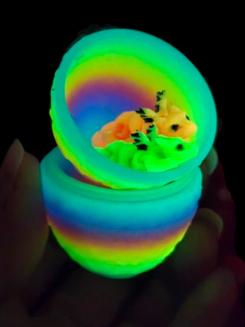 May include: A glowing, multi-colored, rainbow-striped egg-shaped container that is open at the top. Inside the container, two small, detailed, light-colored creatures with dark markings are nestled together, illuminated by a vibrant neon light.
