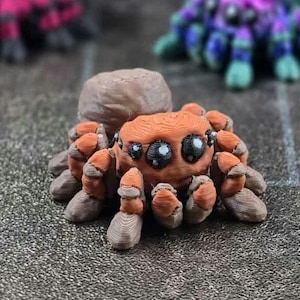 Puede incluir: Figurita detallada de araña naranja y marrón con ojos grandes y expresivos. La araña tiene ocho patas y un cuerpo texturizado, con una cabeza redondeada. Otras figuritas de arañas están borrosas en el fondo.